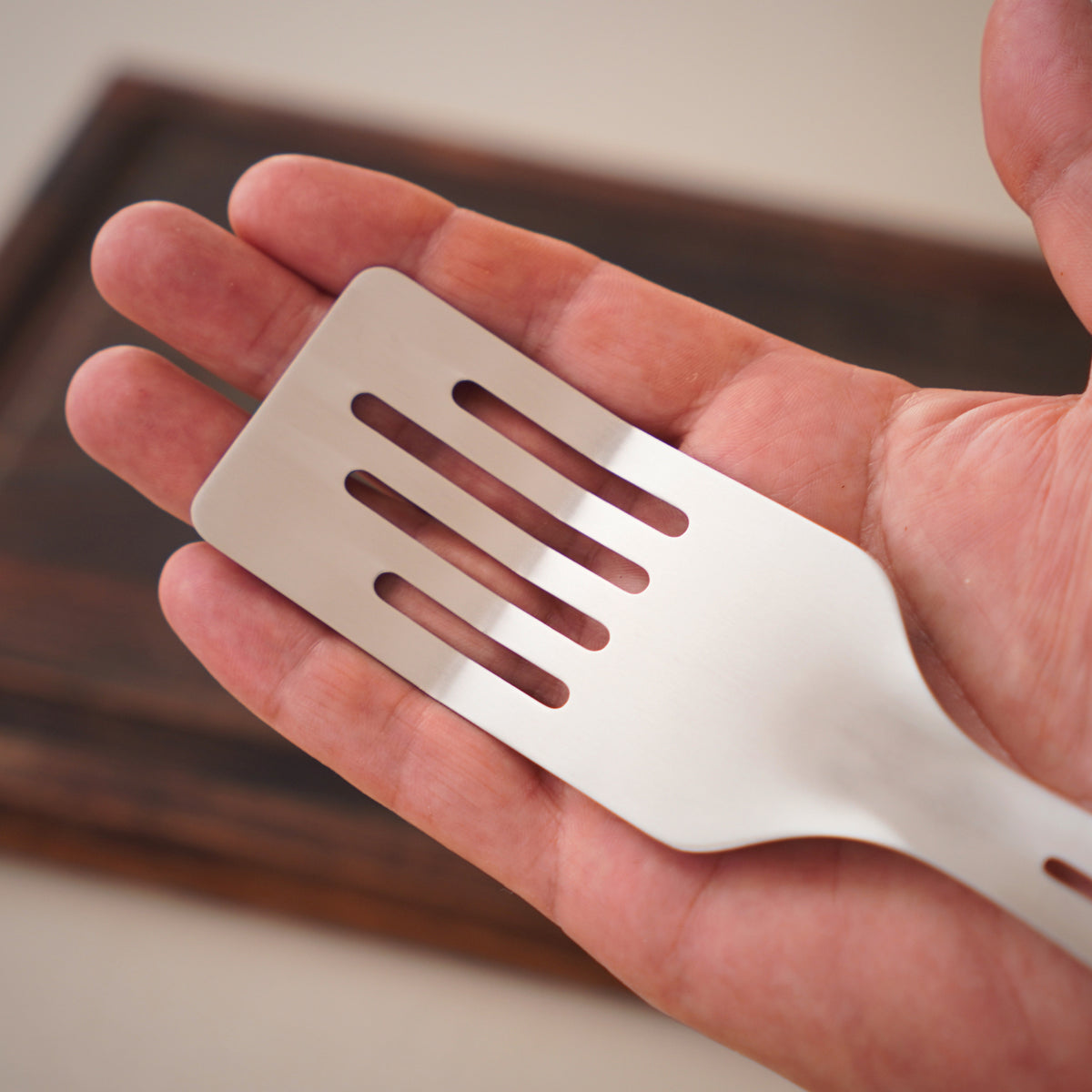 Yahgan Mano one-piece stainless steel spatula held in a hand, showing the finger-shaped slots